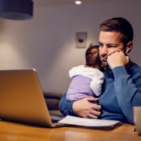 A bored doctors holding his baby girl and having boring online meeting at home.